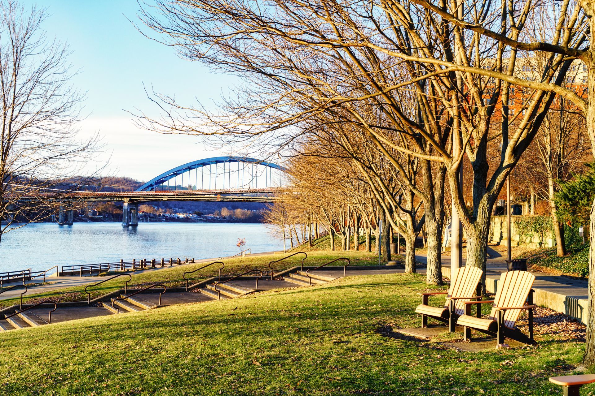 Park with chairs, leafless trees, and river. A blue arched bridge is in the background.