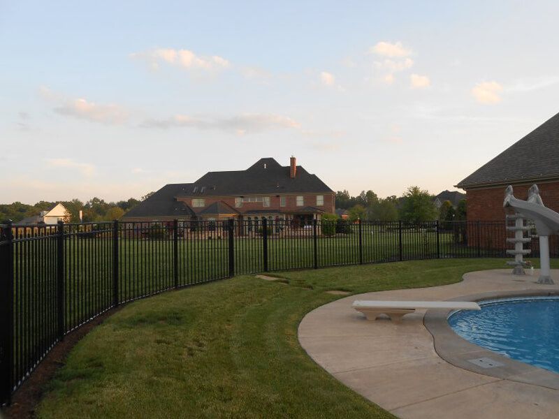 Backyard pool beside a black fence, with a house and grassy lawn at sunset