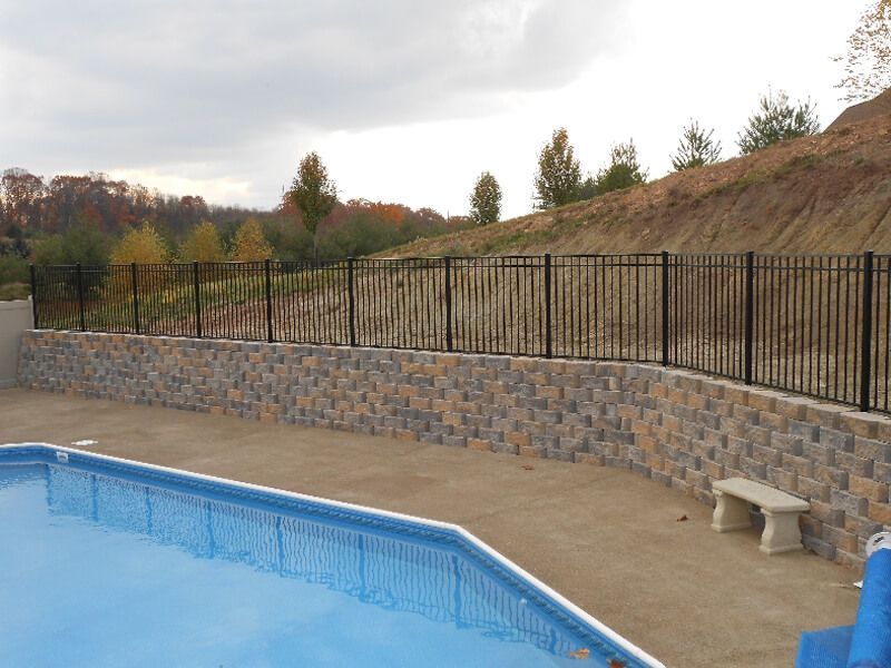 Empty outdoor swimming pool beside a stone wall and black fence on a hillside.
