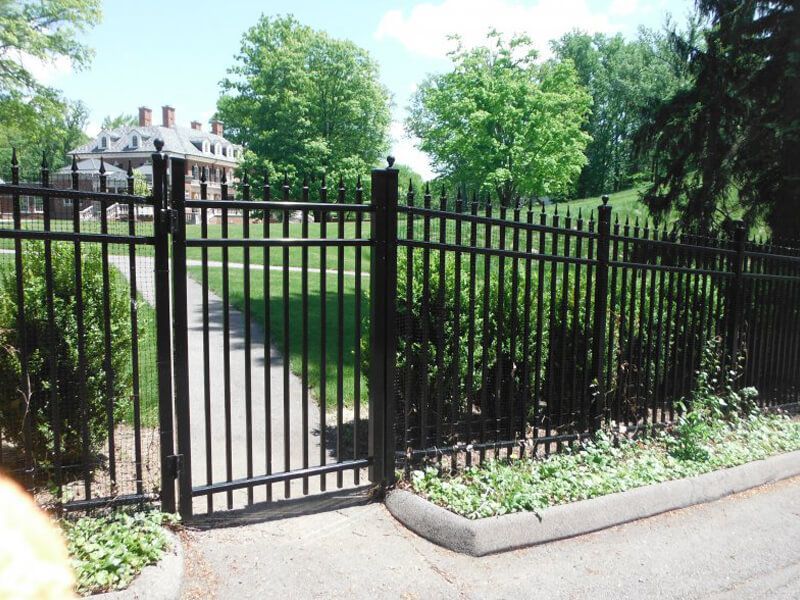 Black metal fence with open gate along a paved path in a green park, with a white building in the background