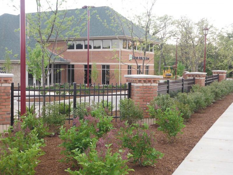 Brick campus building behind black fence, with landscaped flowers and a sidewalk in front.