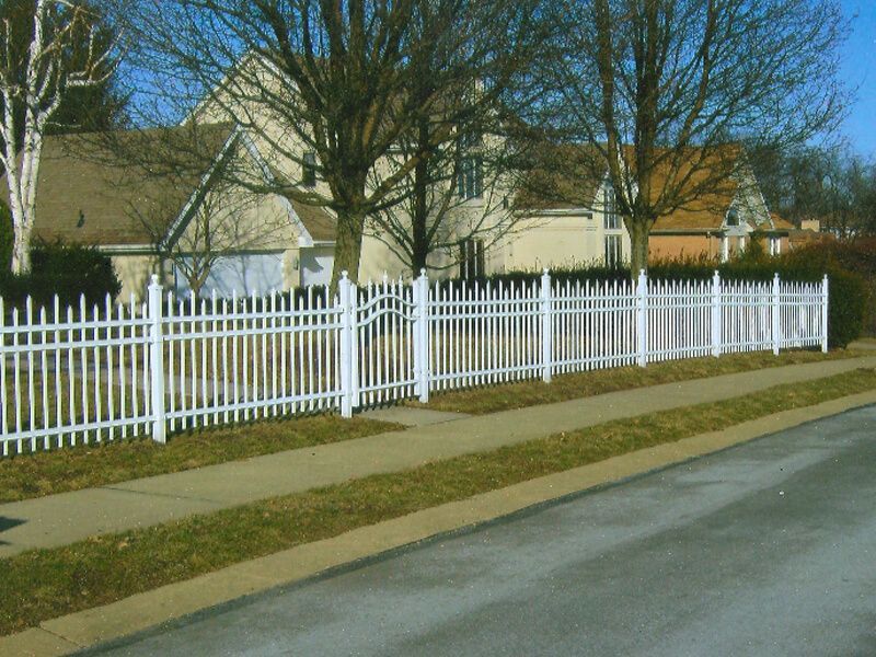 White picket fence along a suburban sidewalk beside houses and bare trees on a sunny day