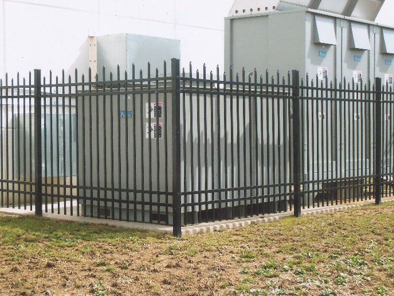 Black metal fence around utility equipment beside a white industrial building.