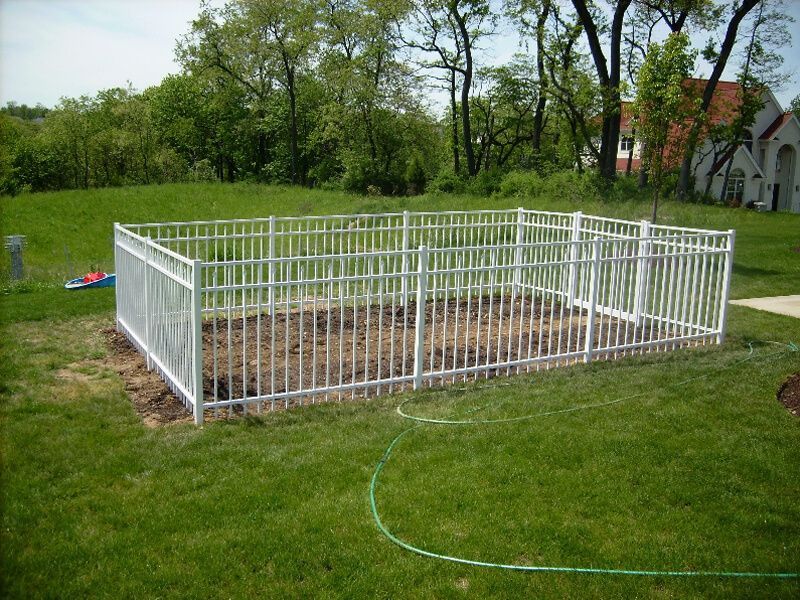 White fenced garden plot with bare soil in a grassy yard, with trees and a house in the background