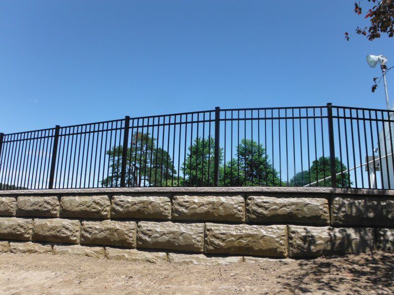 Stone retaining wall with black metal fence under a clear blue sky, with trees in the background