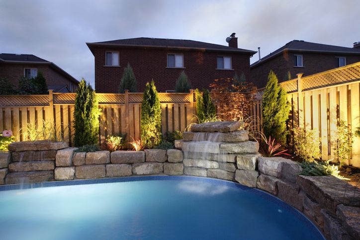Backyard pool with stone retaining wall, lit shrubs, and wooden fence at dusk