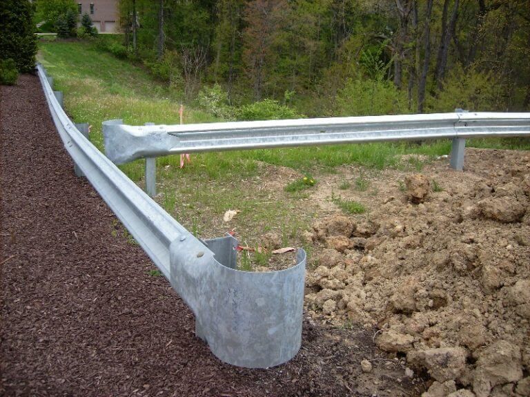 Damaged roadside guardrail with bent metal end near a gravel shoulder and muddy embankment