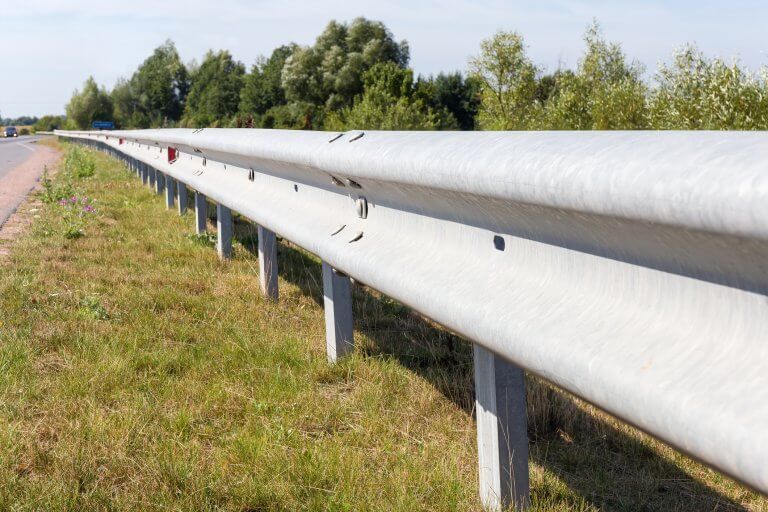 Silver highway guardrail beside a grassy roadside with trees in the background