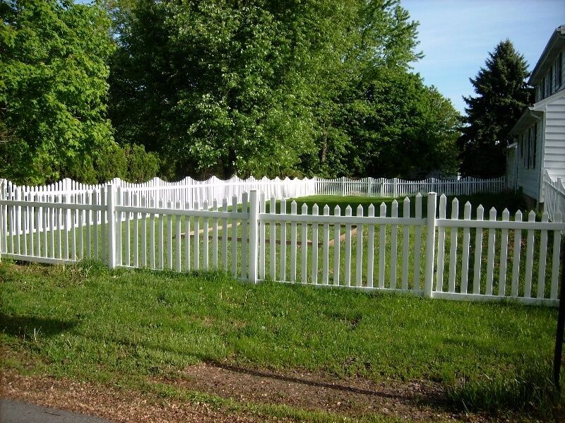 White picket fence in a grassy yard beside a house and tall green trees.