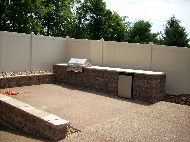 Backyard outdoor kitchen with brick grill, stone counter, and white privacy fence