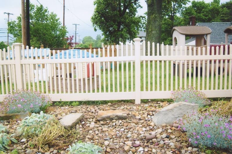 White picket fence behind a rock garden with flowers and trees in a residential yard