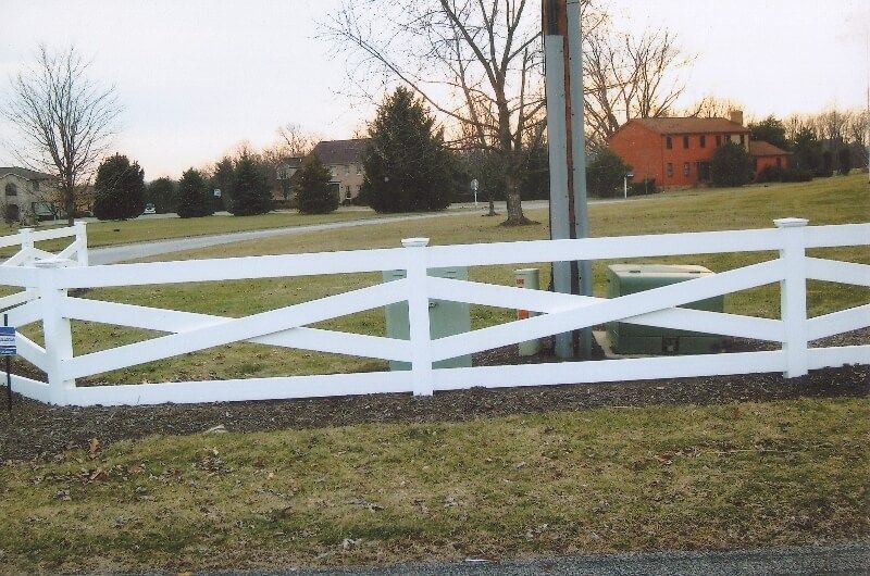 White wooden fence in a grassy field with trees and houses in the background.
