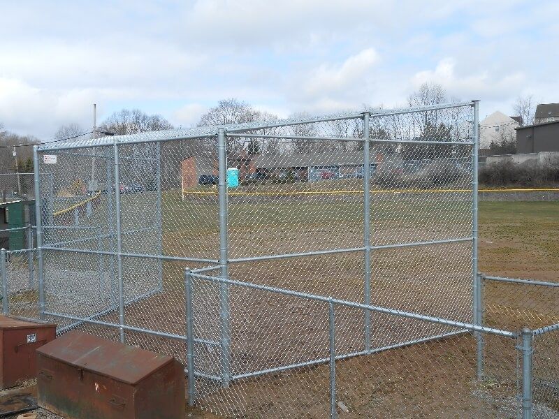 Chain-link sports cage on a grassy field with a small gate and rusty metal boxes in the foreground