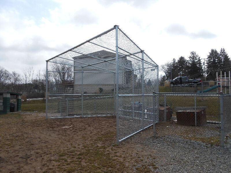 Chain-link fenced enclosure on gravel near trees under a cloudy sky