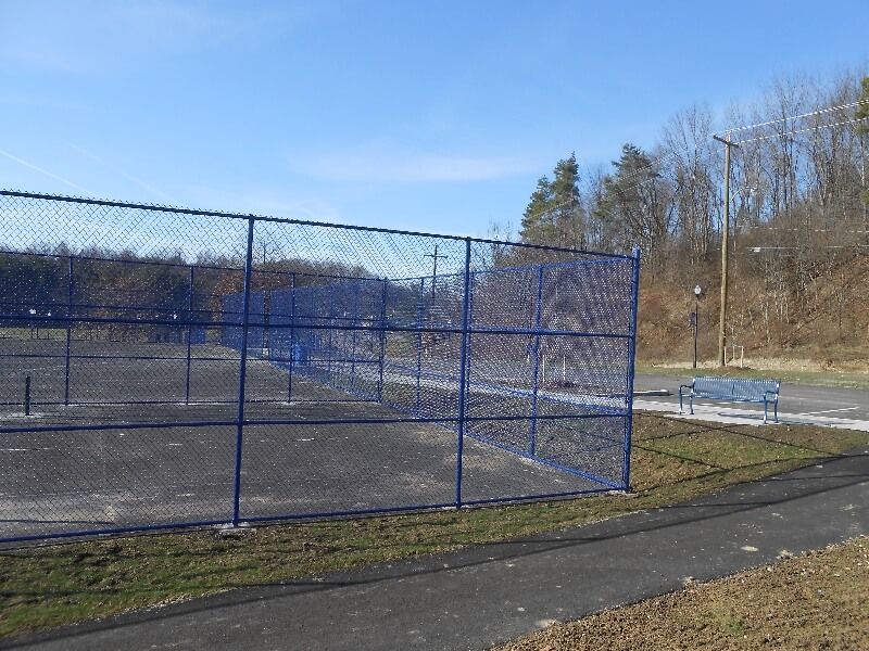 Blue chain-link fence around empty tennis courts on a sunny day, with a bench and trees nearby.