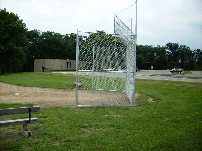 Baseball field backstop and fence beside a dirt infield, with a small building and trees in the background
