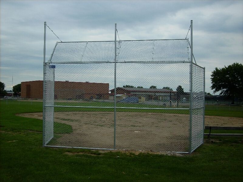 Chain-link batting cage on a grassy field under a cloudy sky