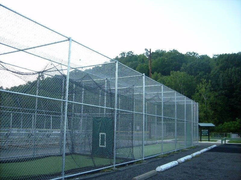 Chain-link fenced sports field with netting, beside a paved path and green trees