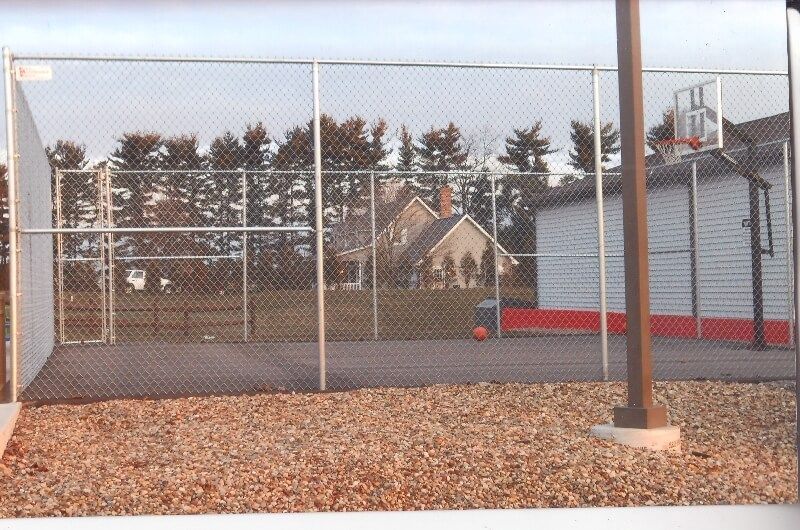 Outdoor basketball court with chain-link fence, hoop, and a small red ball near the baseline.