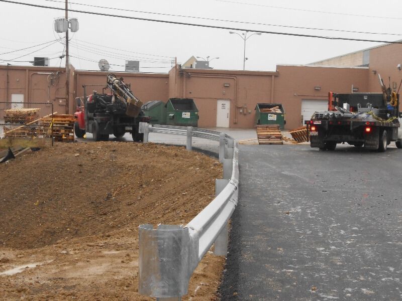 Construction site with trucks and equipment beside a curved metal guardrail on a dirt road