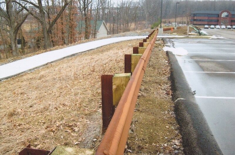 Rust-colored guardrail beside a wet roadside path on a cloudy day, with a parking lot and bare trees nearby