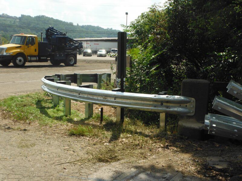 Curved roadside guardrail beside a dirt lot with a yellow truck in the background