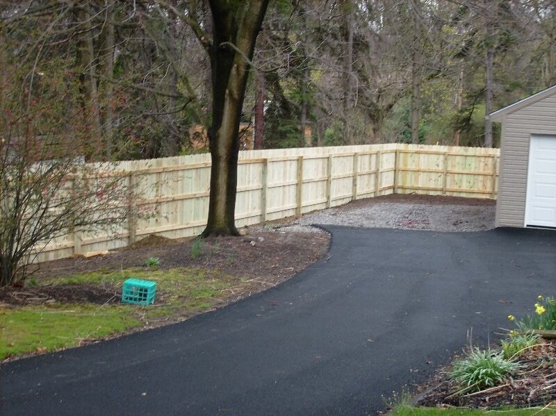 Curved black driveway beside a tan fence, tree, and white garage in a wooded yard