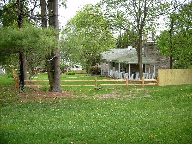 Grassy yard with large trees, a small white house, and a wooden fence in the background