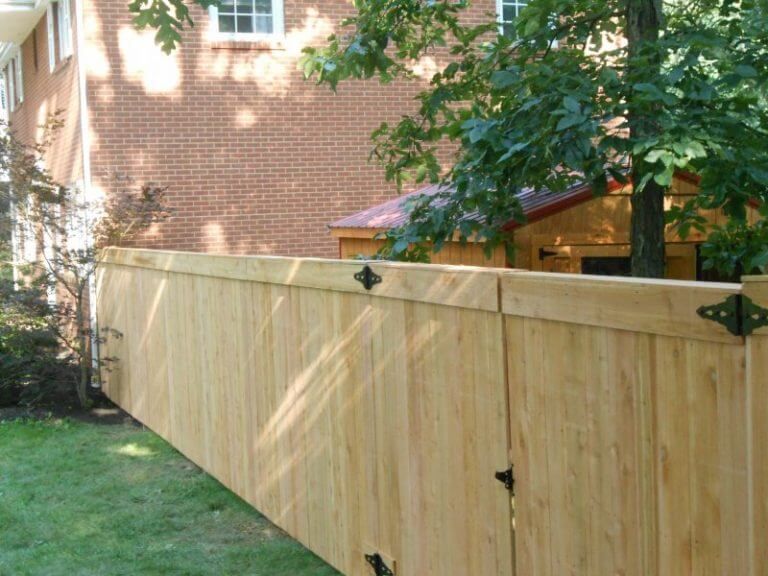 Wooden backyard fence beside a brick house and lawn, with a tree overhead and gate hardware visible