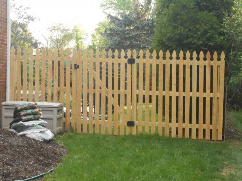 Wooden picket fence with arched tops in a grassy yard, beside stacked stones and trees