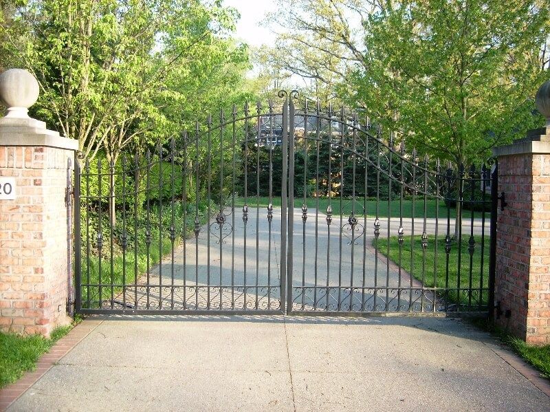 Black metal gate blocking a driveway between brick pillars and trees at the entrance of a property