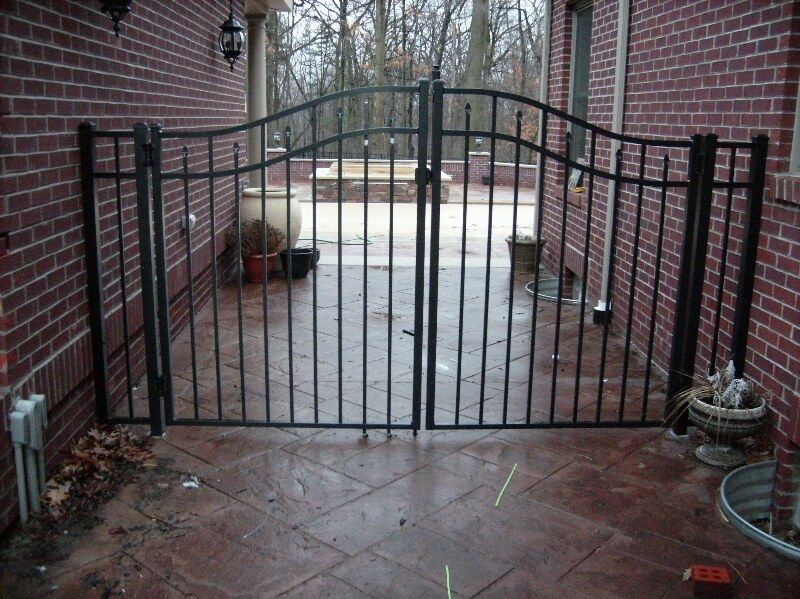 Black metal gate across a narrow brick courtyard with wet tile floor and potted plants.