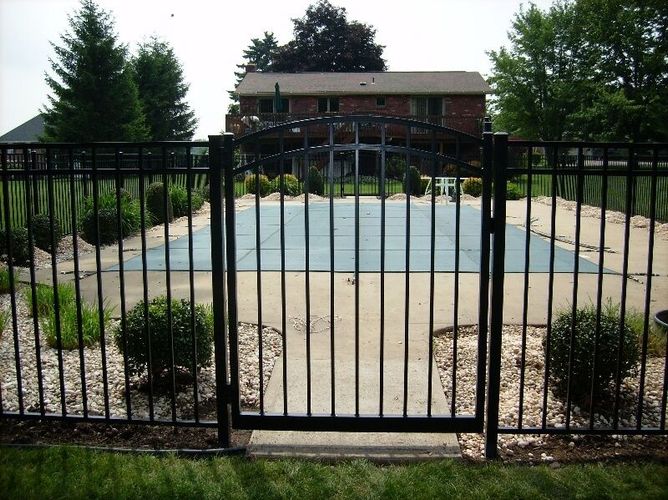 Black metal fence in front of an empty backyard pool with a brick house and trees behind it.