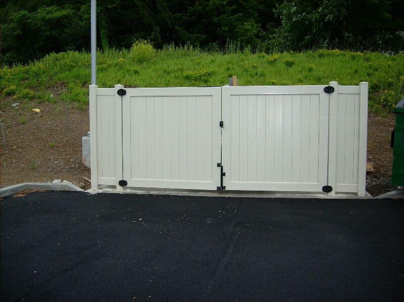 White double driveway gate beside black asphalt, with green foliage behind it.