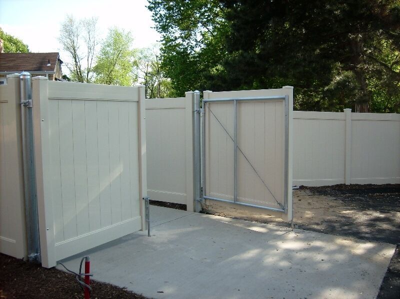 Open white wooden gates in a fenced driveway with concrete ground and trees beyond