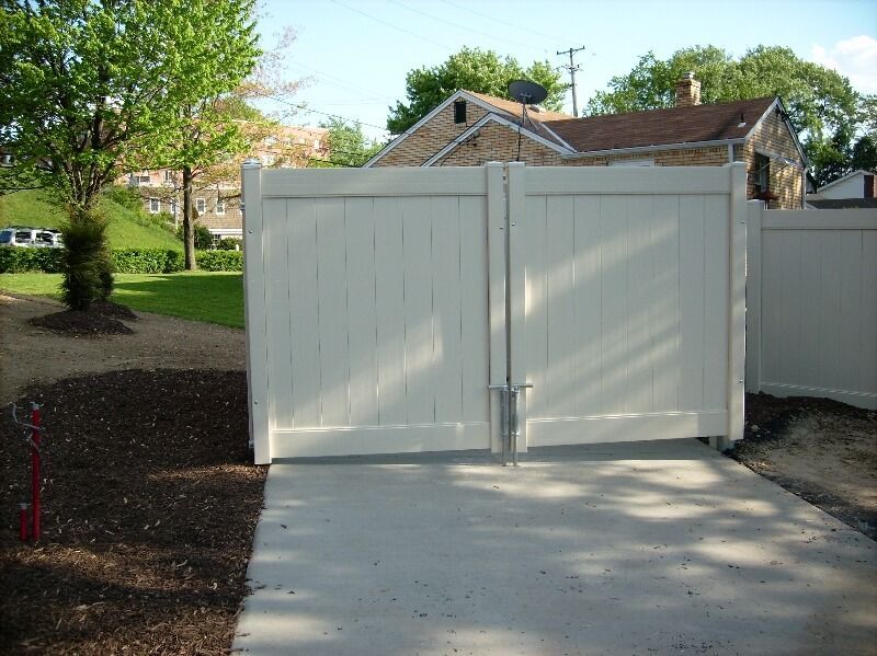 White double gate blocking a driveway, with trees and a house in the background.
