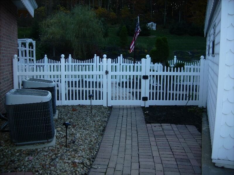 White picket fence and gate at the end of a brick walkway, with a house and yard at dusk.