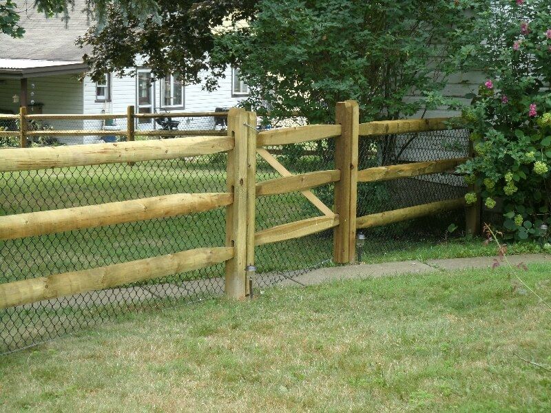Wooden split-rail fence beside a grassy yard with a house and trees in the background