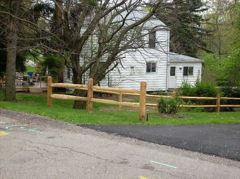 Small white house behind a wooden fence beside a road, surrounded by trees