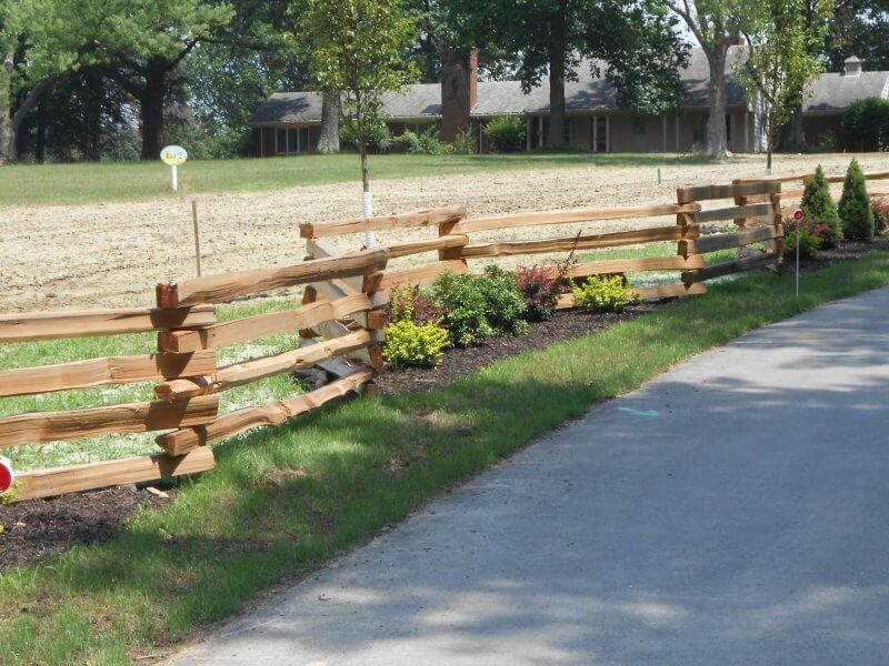 Wooden split-rail fence edging a landscaped grassy strip beside a paved path in a park