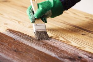 Gloved hand painting a wooden surface with a brush, applying dark stain over light wood.