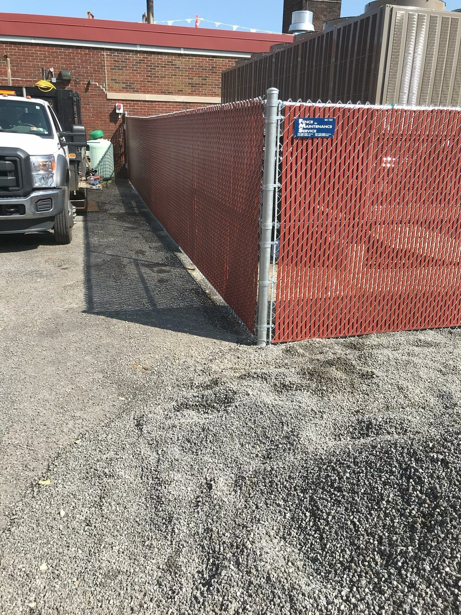 Gravel alley beside a red chain-link fence and brick building, with a parked truck at left.