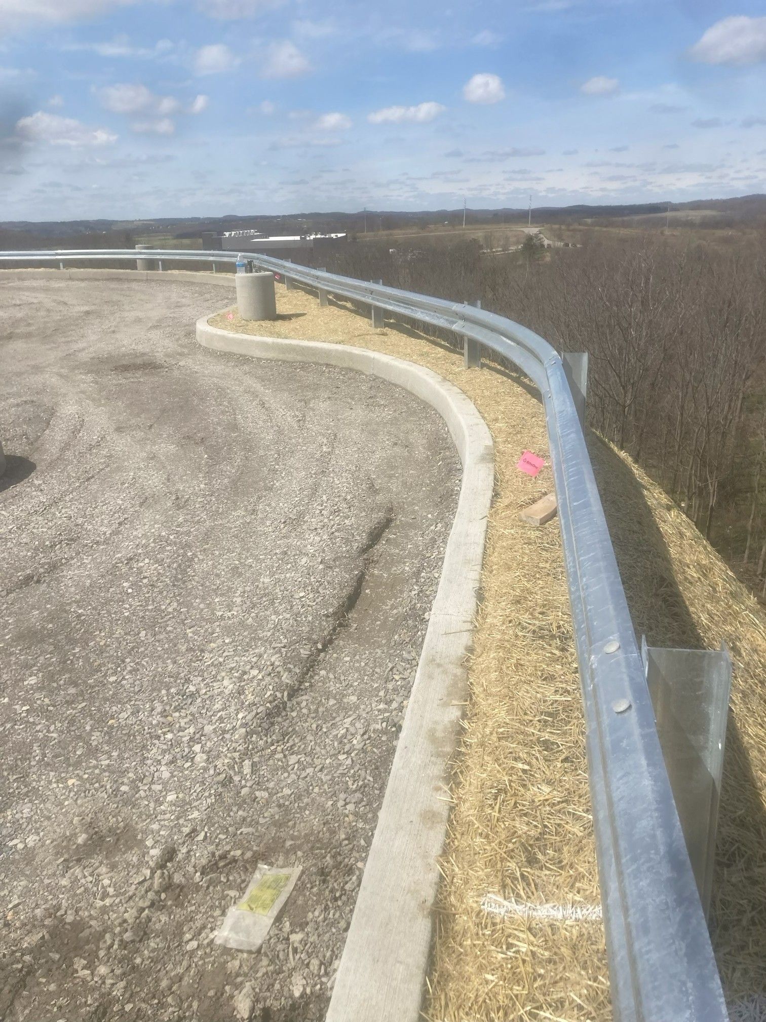 Curving roadside guardrail beside gravel shoulder and open field under cloudy sky