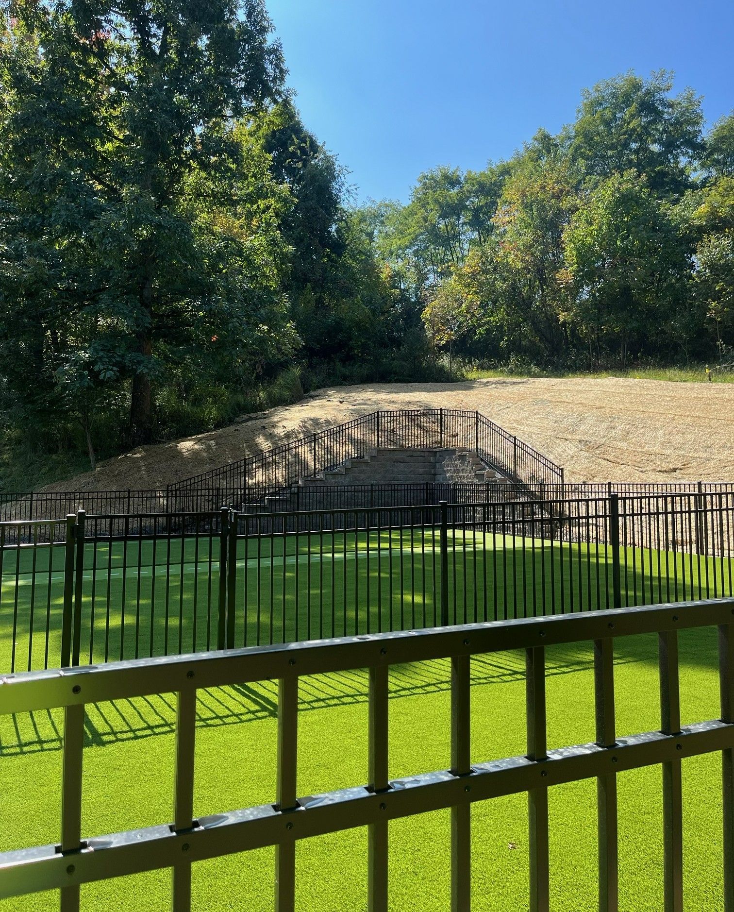 Fenced grassy enclosure with a stone embankment and trees under a blue sky