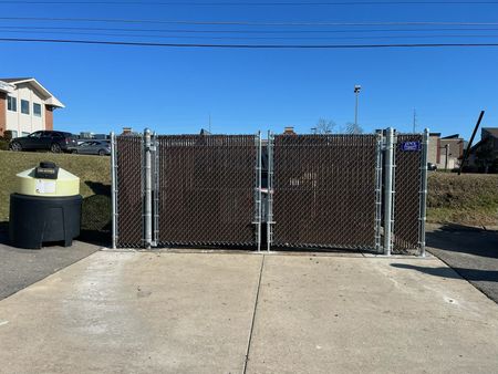 Chain-link gate blocking a concrete driveway, with a trash can on the left and blue sky overhead