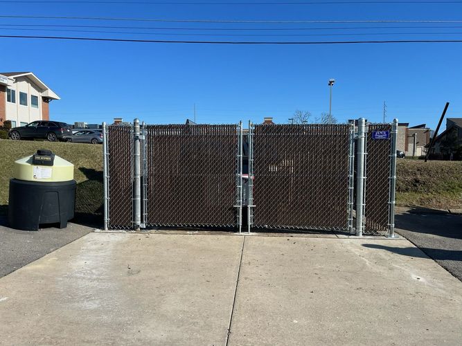 Chain-link gate blocking a driveway, with a concrete path and trash bins under a clear blue sky.