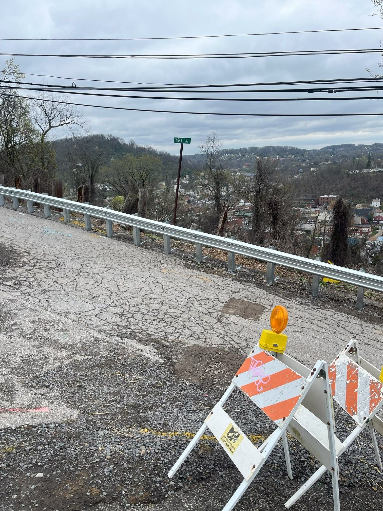 Roadside barrier on a cracked shoulder overlooking a wooded valley under cloudy skies.