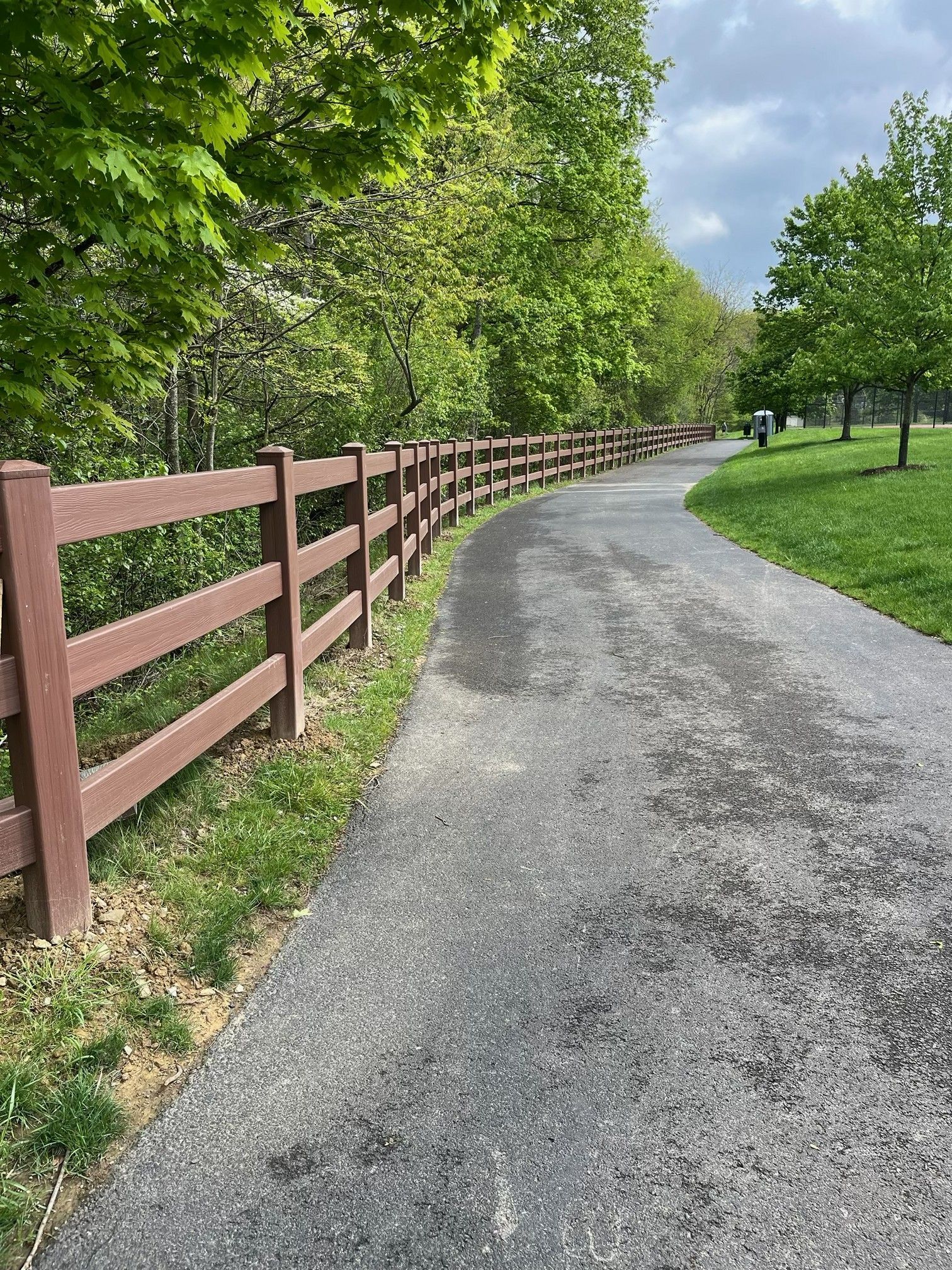 Paved path curving through a green park beside a red wooden fence and trees