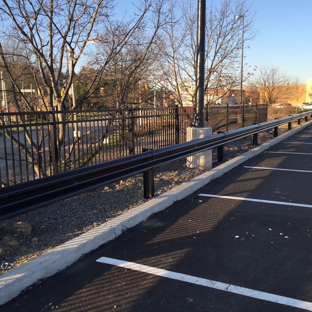 Empty parking lot beside a black fence and road at sunset, with bare trees and long shadows.