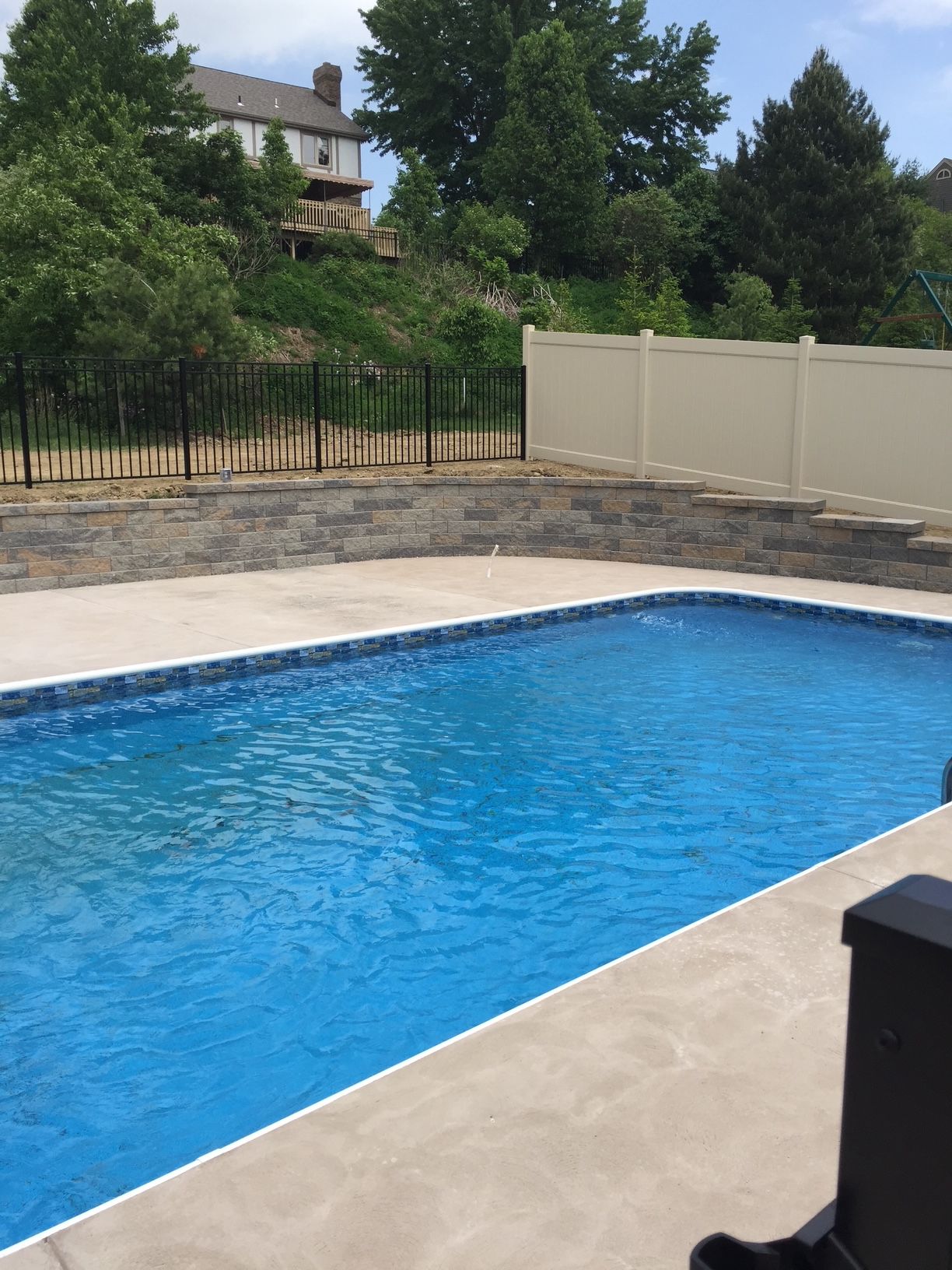 Backyard swimming pool with blue water beside a beige patio and fence, trees and a house in the background